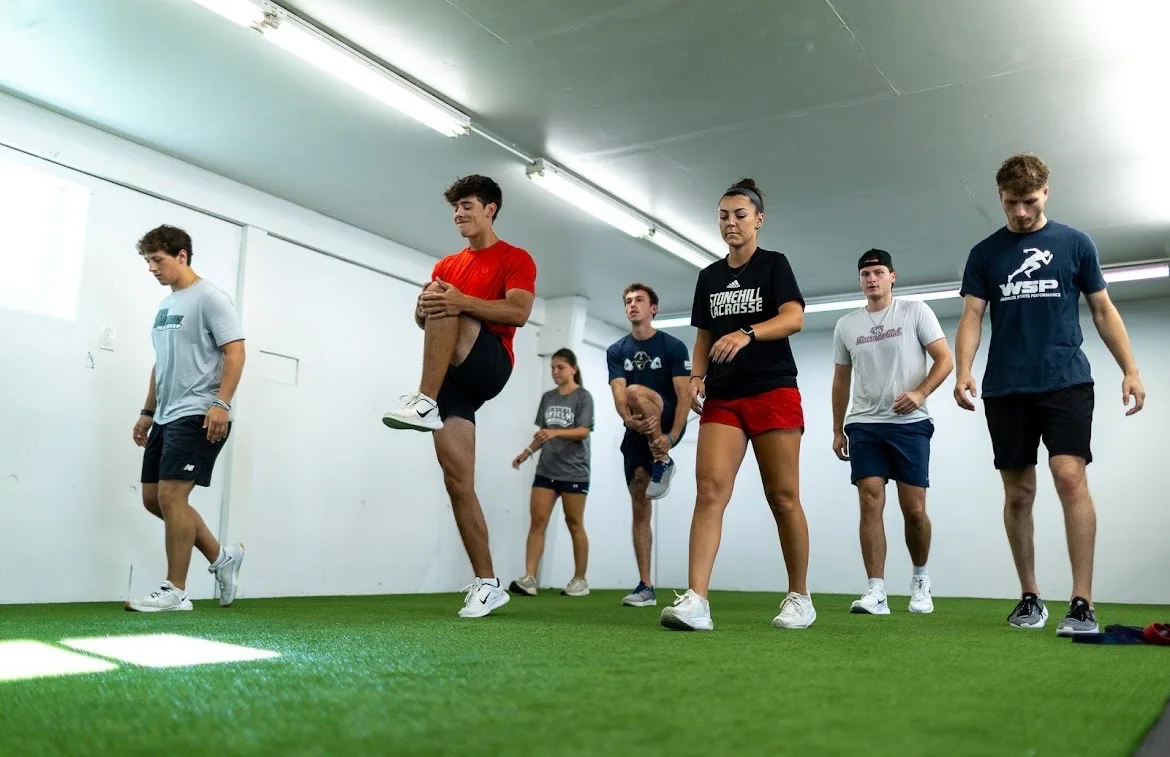 College athletes walking toward the camera while performing knee-hug stretches on the turf inside Warrior Sports Performance